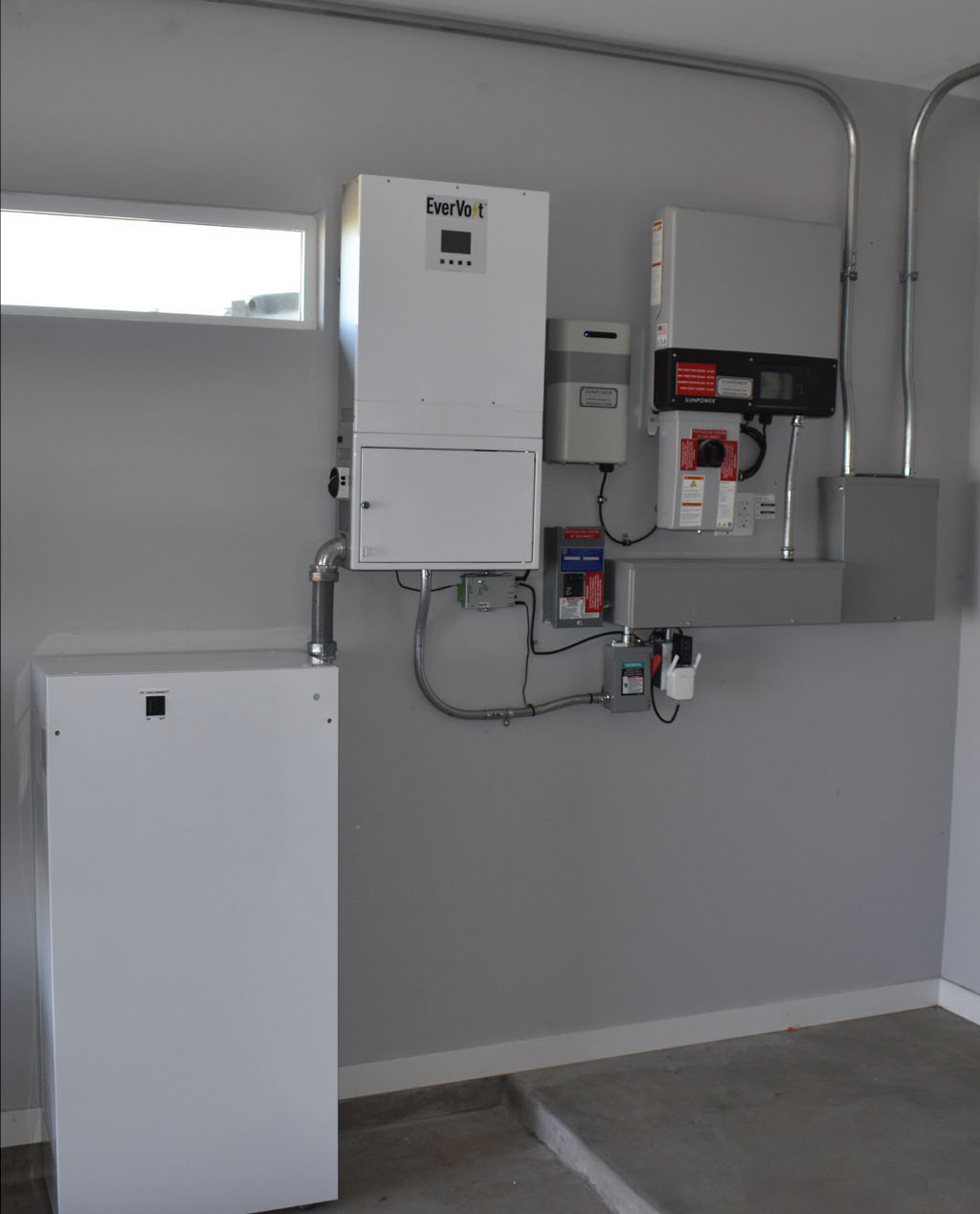 Photo 6. Battery backed up system by EverVolt (Panasonic USA). White battery enclosure on the left. White multimode inverter in the center. AC coupled to PV system with Gray/black SMA Sunny Boy utility-interactive inverter to the right. Courtesy of John Wiles